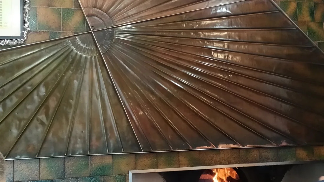 La cappa di rame a ventaglio sopra il forno, le piastrelle verdi e ocra della parete e le due lampade a sospensione.
