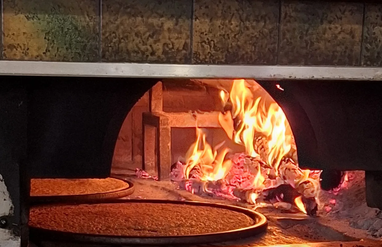 La bocca del forno a legna accesa: due teglie tonde di cecina appoggiate sul fondo, la fiamma e la brace sulla destra.