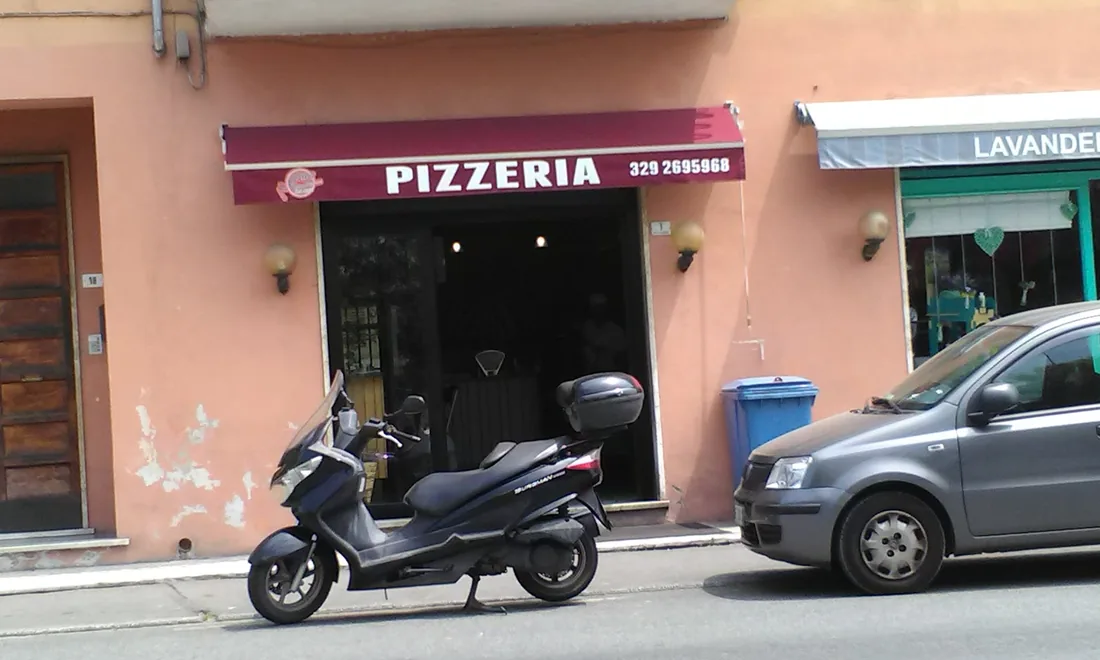 L'ingresso della pizzeria in Piazza IV Novembre: tenda bordeaux con la scritta PIZZERIA e il numero di telefono, facciata rosa e porta aperta.
